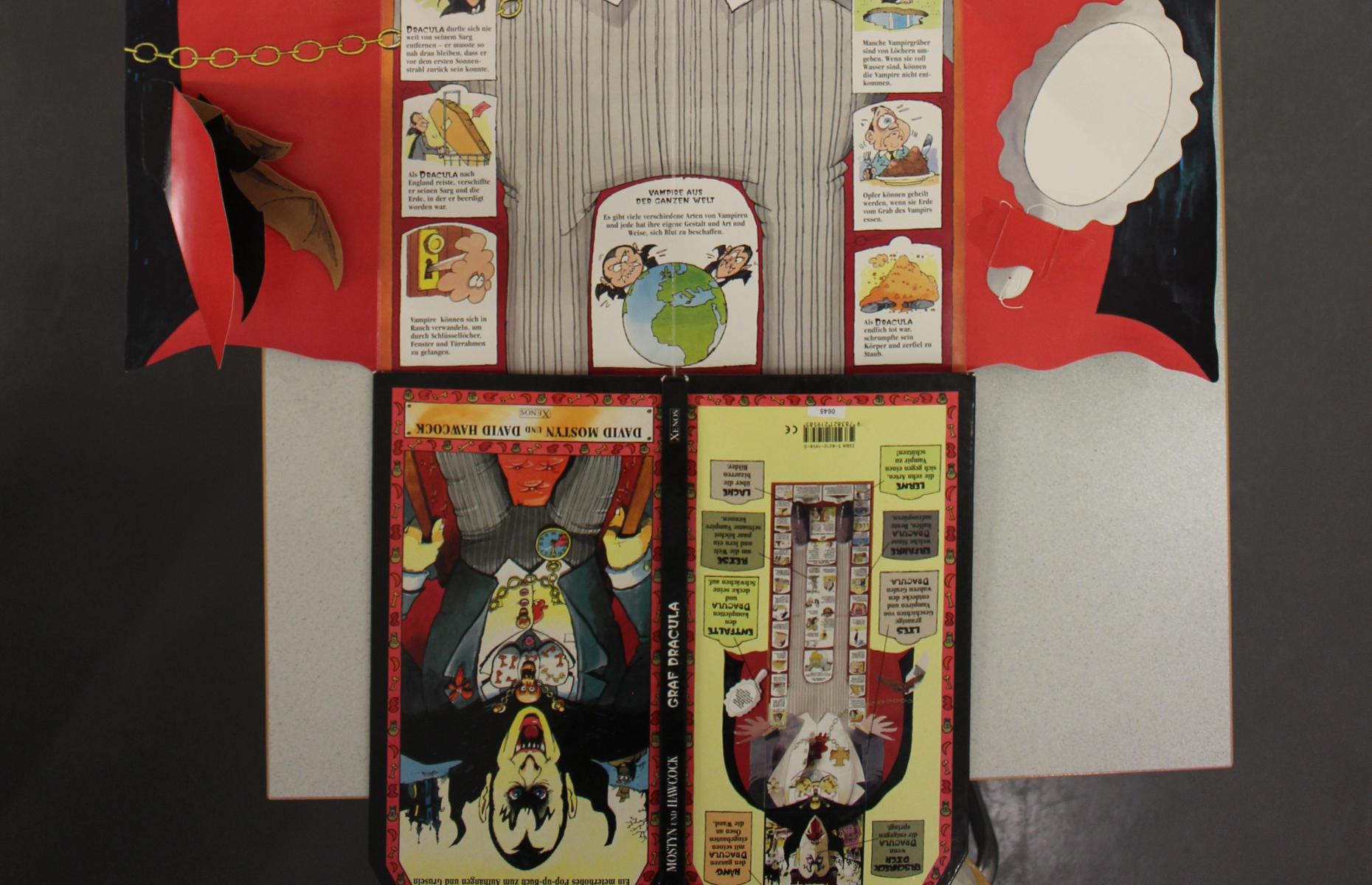 Unterer Teil des vollständig aufgeklappten Pop-up-Buch &quot;Graf Dracula&quot;. Zu sehen sind der untere Teil von Graf Draculas Umhang, die Texte des Buches sowie die Vorder- und Rückseite des Buches.